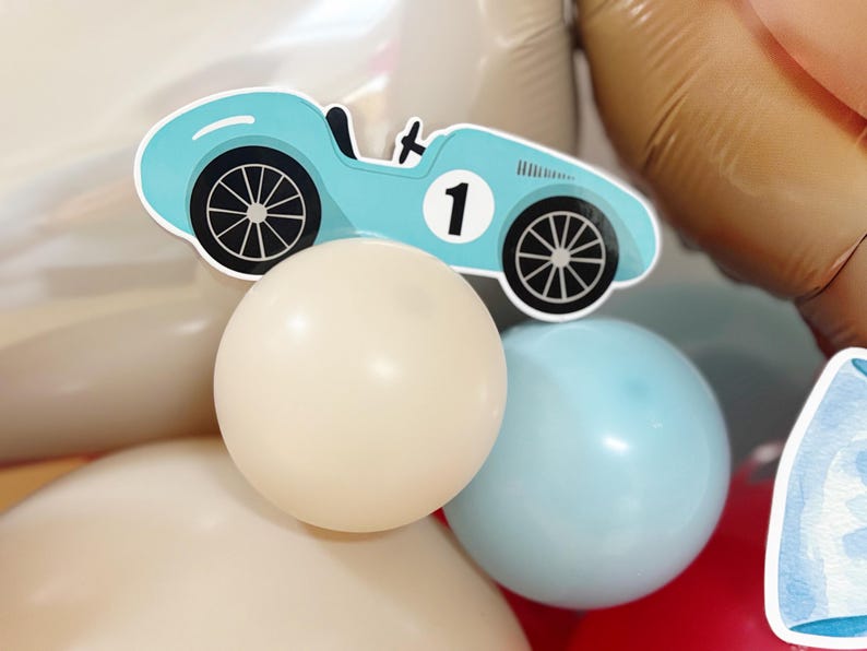 May include: A light blue race car cutout with the number 1, placed above cream and light blue balloons. The car has black wheels and a black steering wheel. The balloons are part of a party decoration.