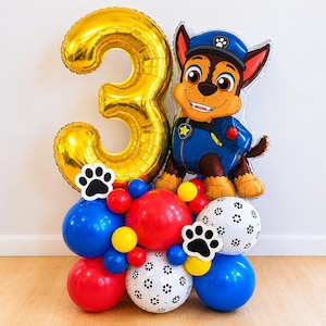 May include: A festive balloon arrangement featuring a gold number 3 balloon and a Chase from Paw Patrol character balloon. The base includes red, blue, yellow, and white balloons, creating a colorful display for a birthday celebration.