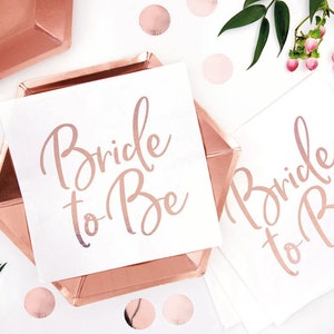 May include: Rose gold paper plates and napkins with the text "Bride to Be" in a cursive font. The plates are hexagonal and surrounded by rose gold confetti and greenery, perfect for a bridal shower.