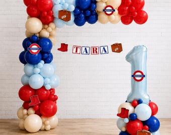Könnte beinhalten: Ein Ballonbogen und eine Ballon-Säule in Form der Zahl Eins aus roten, blauen, hellblauen und cremefarbenen Ballons. Das Set enthält Ausschnitte von roten Stiefeln, braunen Koffern und runden Schildern mit dem Text Paddington. Ein Banner zeigt TARA.