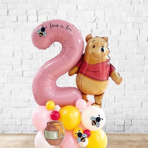 以下が含まれることがあります： 大きなピンク色の数字2のバルーン、テキスト「Tara is Two」、ハチのイラストが特徴の、お祝いのバルーンディスプレイ。くまのプーさんのバルーンが数字の隣に配置されています。ピンク、黄色、赤のバルーン、ハチミツポットとハチの切り抜きが含まれています。