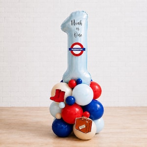 Puede incluir: Un globo de aluminio azul claro con forma de número uno, el texto Noah is One y un logo de Paddington rojo y azul. La base es un conjunto de globos rojos, blancos y azules decorados con botas rojas y un gráfico de maleta marrón.