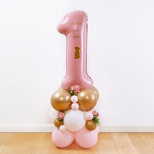Könnte beinhalten: Ein rosa Ballon in Form der Zahl 1, mit dem Namen "Amelia" und dem Wort "One" sowie einer goldenen Hasenillustration. Er steht auf einer Ansammlung goldener, weißer und rosa Ballons, die mit kleinen Blumengestecken verziert sind.