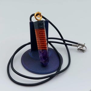 Puede incluir: Un collar con colgante de cristal morado envuelto en alambre de cobre, con un cordón negro y un cierre dorado. El colgante se exhibe sobre un soporte azul oscuro sobre un fondo blanco. El collar es una pieza de joyería única.