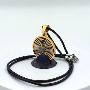 Könnte beinhalten: Goldfarbene Anhänger-Halskette mit einem kreisförmigen Design und einem schwarzen Spiralmuster. Der Anhänger hängt an einer schwarzen Kordelkette mit einem Verschluss. Die Halskette wird auf einem kleinen, dunkelblauen Ständer präsentiert.