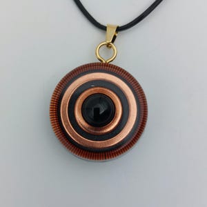 May include: A pendant necklace featuring a circular design with concentric rings of copper and black, centered with a black stone. The pendant hangs from a black cord with a gold-colored bail.