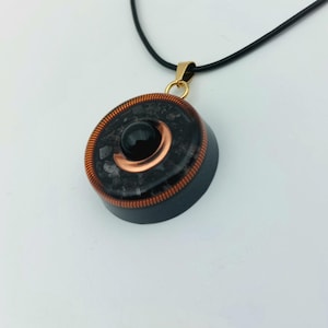 Puede incluir: Un collar con colgante negro con un diseño circular. El colgante presenta un centro de piedra negra, detalles en cobre y una fianza dorada. El cordón del collar es negro.