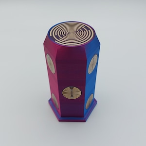 Puede incluir: Un recipiente hexagonal, multicolor, decorativo con una tapa dorada y un patrón de espiral. El recipiente está hecho de un material liso y brillante y tiene un diseño moderno.