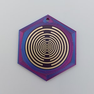 Puede incluir: Un colgante hexagonal con un diseño de círculos concéntricos en oro sobre un fondo morado y azul.