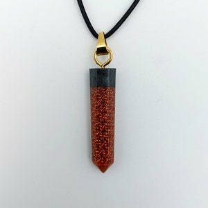 May include: A gold pendant necklace with a black and copper-colored resin pendant. The pendant is shaped like a long, thin point.