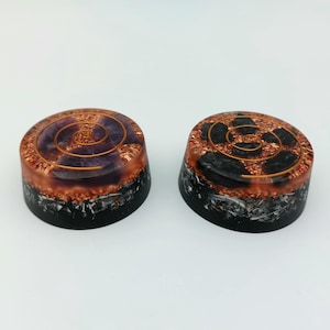 Op de afbeelding: Twee ronde, zwarte en koperkleurige kristallen orgone-piramides met een spiraalvormig ontwerp. Eén piramide heeft een paarse kern en de andere een zwarte kern.