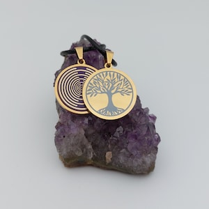 May include: Two gold pendants on a black cord, one with a purple spiral design and the other with a silver tree of life design, displayed on a bed of amethyst crystals.