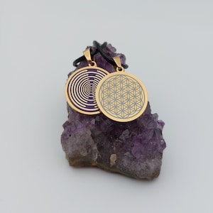 May include: Two gold pendants on black cords, one with a purple and white spiral design and the other with a gold flower of life design, displayed on a bed of amethyst crystals.