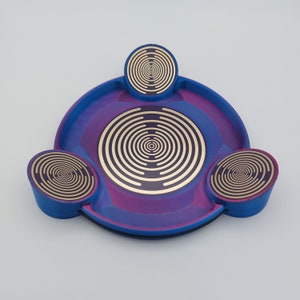 Puede incluir: Una bandeja circular azul y morada con tres compartimentos circulares más pequeños. Cada compartimento tiene un diseño de círculo concéntrico dorado.