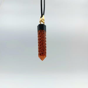 Puede incluir: Un colgante de cristal de color cobre con una parte superior negra, que cuelga de un cord&oacute;n negro. El colgante tiene forma de l&aacute;grima puntiaguda y alargada.