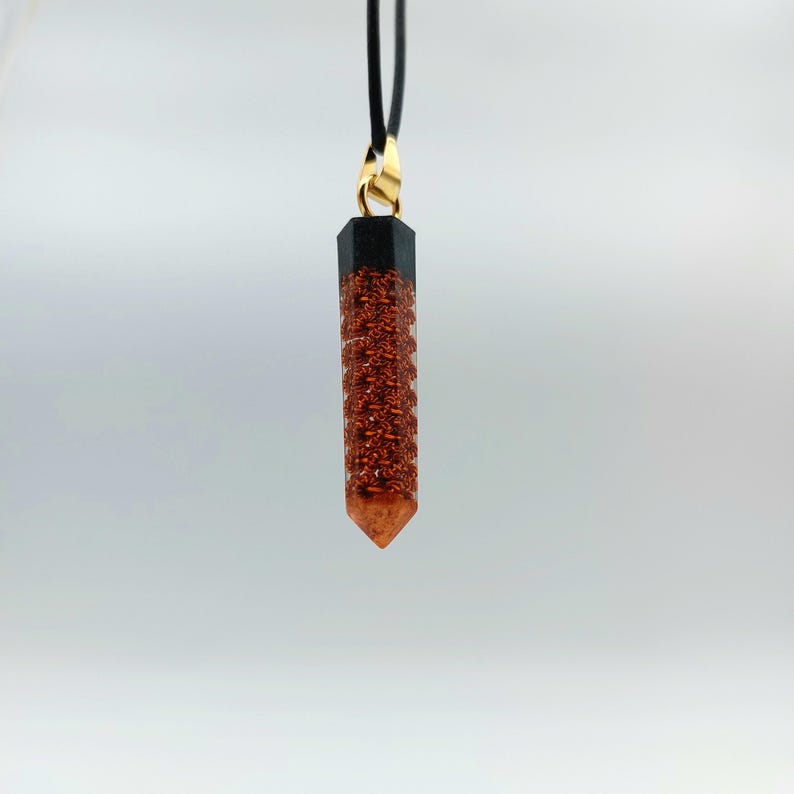 Puede incluir: Un colgante de cristal de color cobre con una parte superior negra, que cuelga de un cord&oacute;n negro. El colgante tiene forma de l&aacute;grima puntiaguda y alargada.