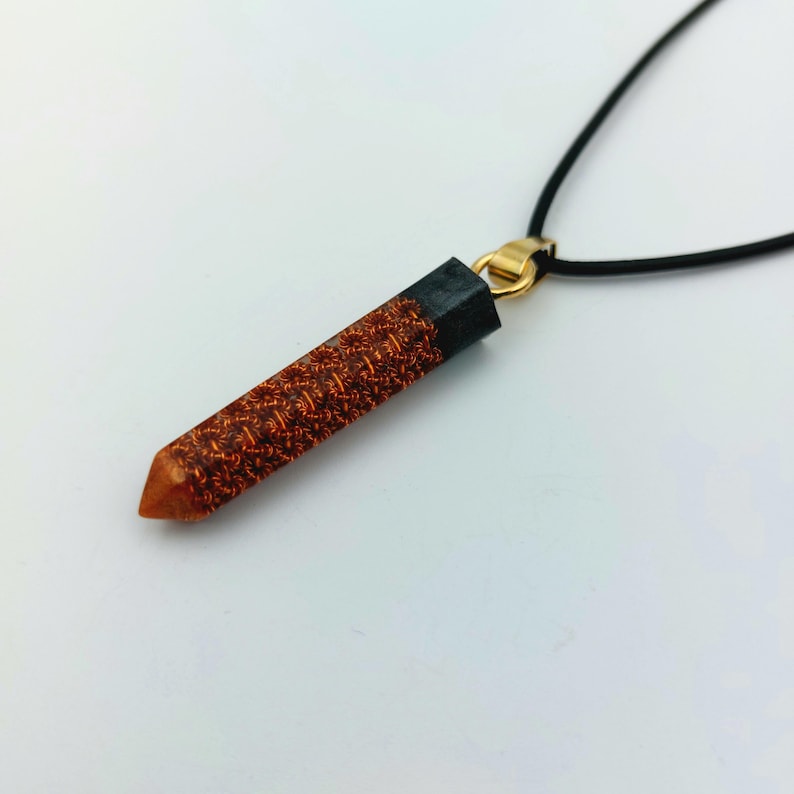 Puede incluir: Un collar con un colgante largo y delgado hecho de resina. El colgante es de color marr&oacute;n oscuro con un patr&oacute;n de color cobre. El colgante est&aacute; unido a un cord&oacute;n negro.