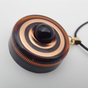 May include: A pendant with a black gemstone center and copper spiral design. The pendant is round with a dark brown border and a gold-colored clasp. The necklace cord is black.
