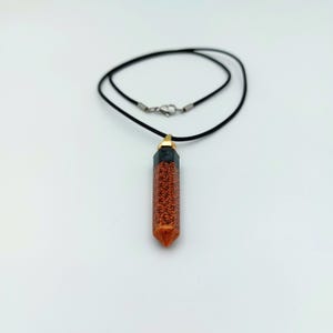Puede incluir: Un collar de cord&oacute;n negro con un colgante de cristal puntiagudo relleno de purpurina naranja con punta dorada.