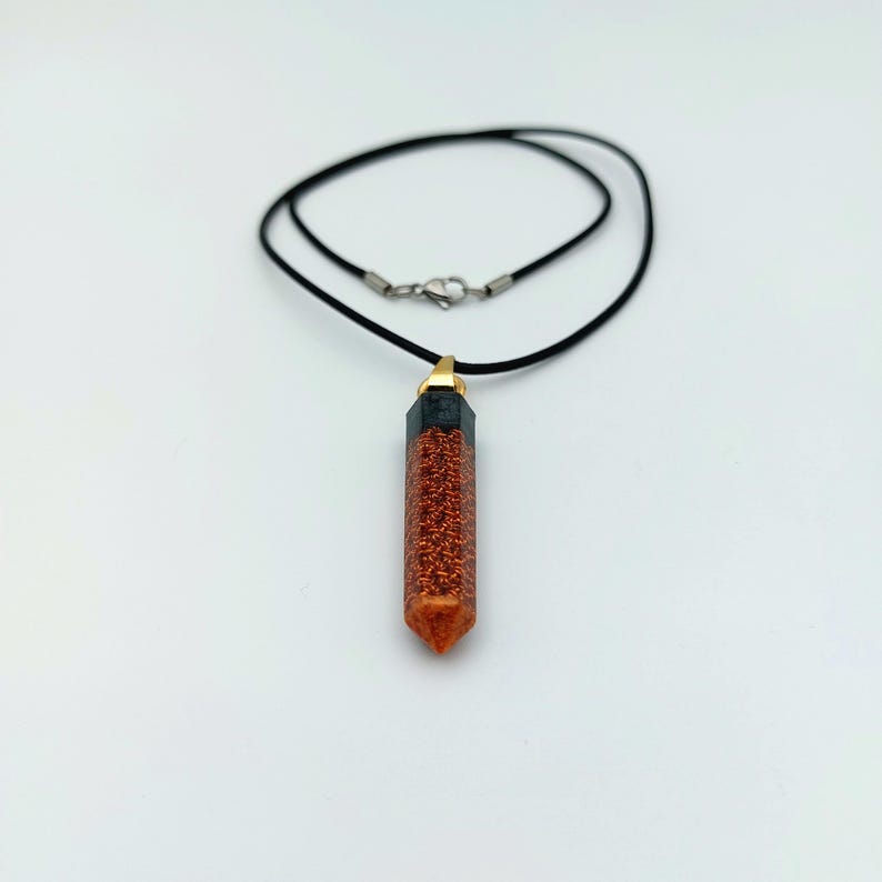 Puede incluir: Un collar de cord&oacute;n negro con un colgante de cristal puntiagudo relleno de purpurina naranja con punta dorada.