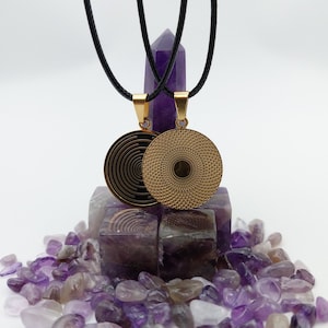 Puede incluir: Dos collares con colgantes dorados con cordones negros. Un colgante presenta un diseño en espiral, el otro un patrón texturizado. Los colgantes cuelgan sobre un fondo de cristales de amatista y un cristal alto y púrpura.