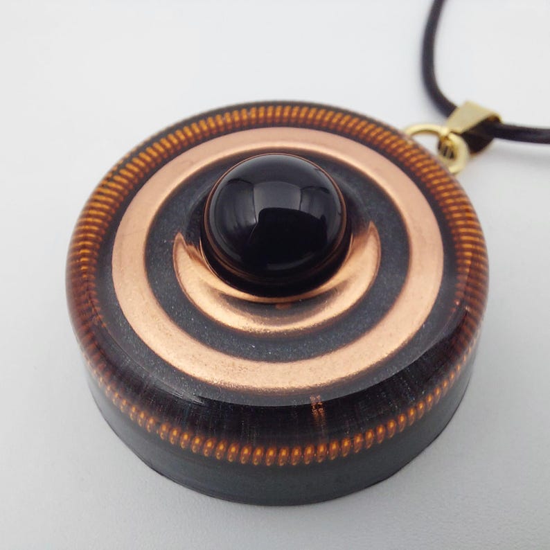 May include: A round pendant with a black gemstone center and copper-colored concentric circles. The pendant is attached to a dark brown cord with a gold-colored clasp. The outer edge has a textured, copper-colored design.