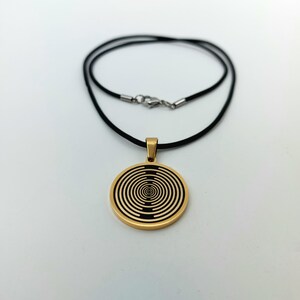 May include: A gold pendant necklace with a black and gold circular design. The pendant features a concentric circle pattern. The necklace is on a black cord.
