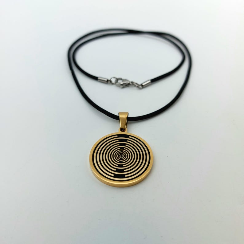 May include: A gold pendant necklace with a black and gold circular design. The pendant features a concentric circle pattern. The necklace is on a black cord.