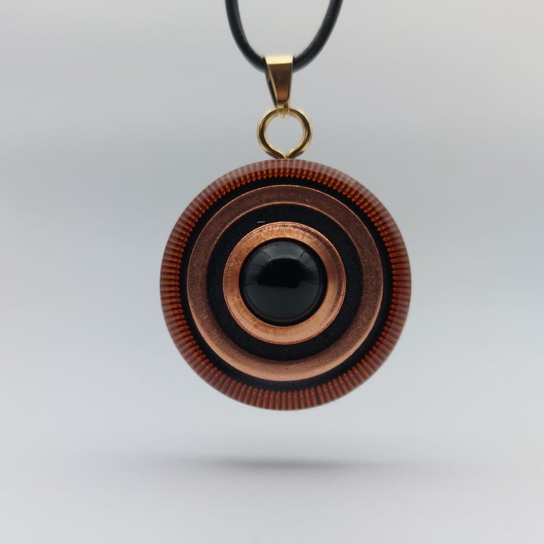 May include: A round pendant necklace with a black cord. The pendant features concentric circles in copper and black, with a black stone center. The pendant is suspended from a gold-colored bail.