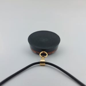 May include: A black pendant with a gold-colored bail and a black cord. The pendant has a circular, flat top and a copper-colored band around the side. The pendant is likely a piece of jewelry.