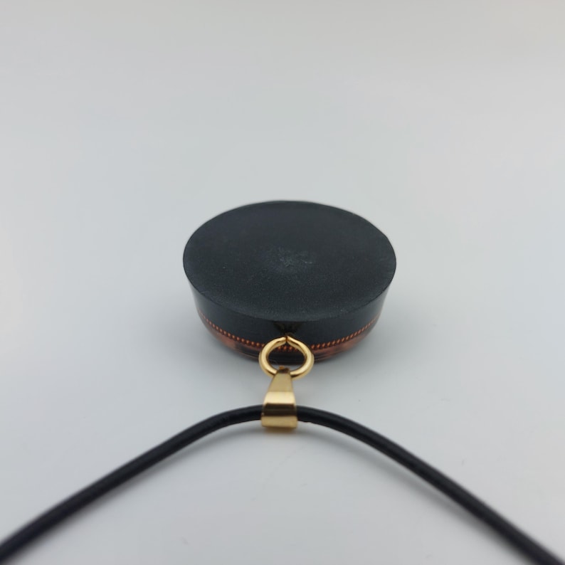 May include: A black pendant with a gold-colored bail and a black cord. The pendant has a circular, flat top and a copper-colored band around the side. The pendant is likely a piece of jewelry.