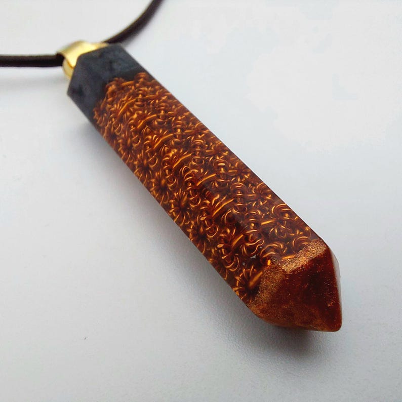 Puede incluir: Un collar con un colgante de resina marr&oacute;n y dorado, largo y delgado, con una cadena dorada y negra. El colgante tiene un patr&oacute;n detallado e intrincado.