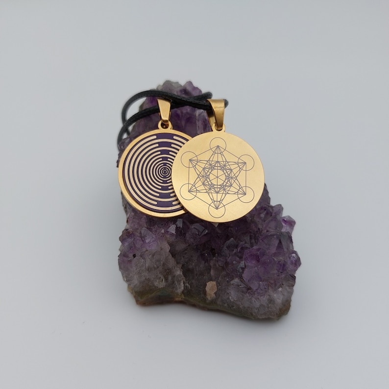 May include: Two gold pendants on a black cord, resting on a bed of amethyst crystals. One pendant features a geometric design, the other a swirling pattern.