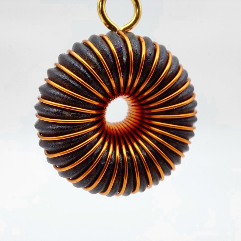May include: A copper and black wire coil pendant with a gold loop at the top. The coil is wound in a spiral pattern and has a circular opening in the center.
