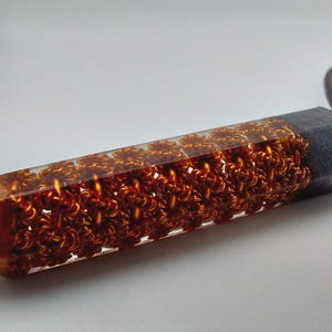 Puede incluir: Un colgante de resina transparente con un dise&ntilde;o de alambre de cobre en su interior. El alambre est&aacute; dispuesto en un patr&oacute;n repetitivo de peque&ntilde;os c&iacute;rculos.