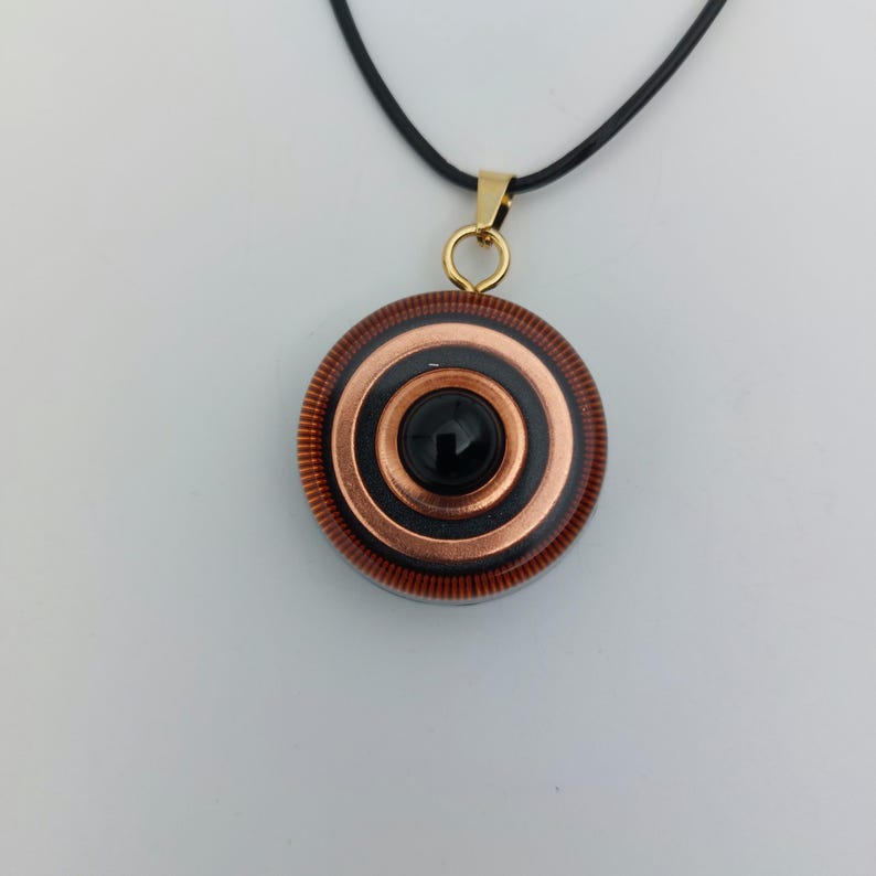 May include: A pendant necklace featuring a circular design with concentric rings of copper and black, centered with a black stone. The pendant hangs from a black cord with a gold-colored bail.