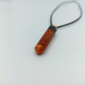 Puede incluir: Un colgante de madera marr&oacute;n y negro con forma geom&eacute;trica que cuelga de un collar de cord&oacute;n negro.