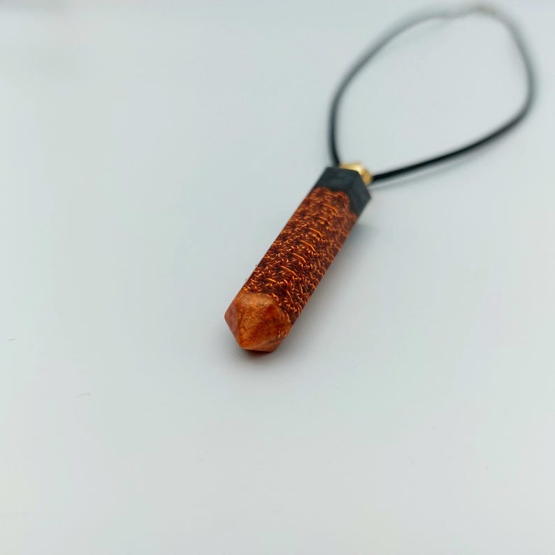 Puede incluir: Un colgante de madera marr&oacute;n y negro con forma geom&eacute;trica que cuelga de un collar de cord&oacute;n negro.
