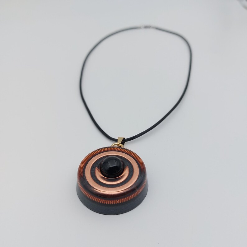 May include: A pendant necklace featuring a circular pendant with concentric copper and dark wood rings. A small, black, round stone is centered on the pendant. The necklace has a black cord and a gold-colored clasp.