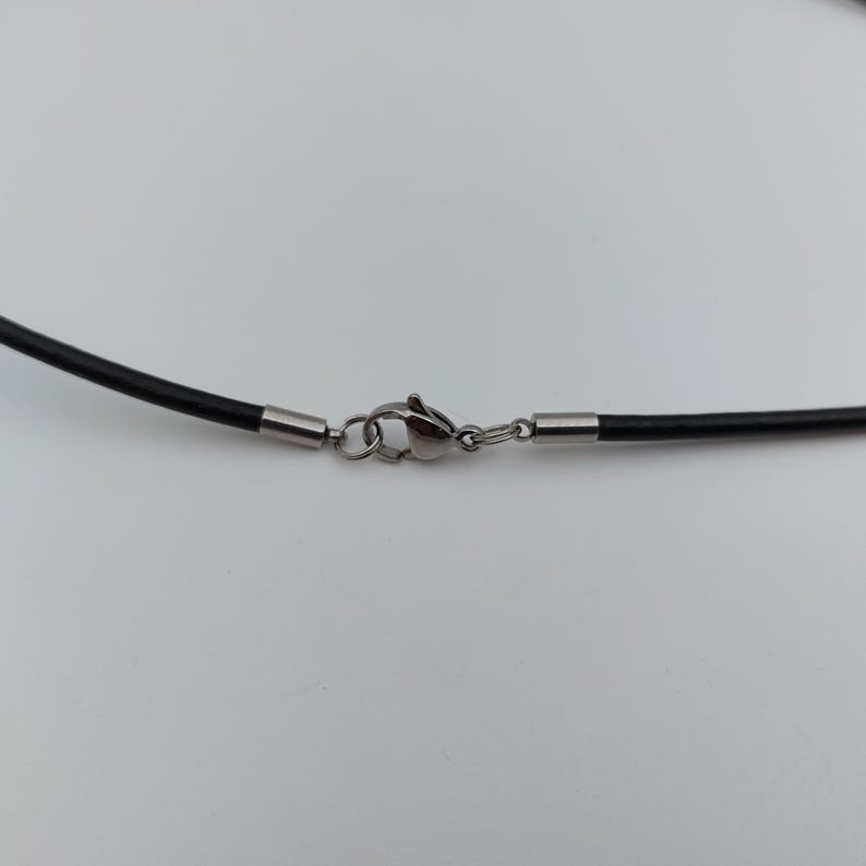 May include: Close-up of a black necklace cord with a silver-colored clasp. The cord is smooth and appears to be made of a flexible material. The clasp is a lobster claw style, with a small ring to attach to the other end of the necklace.