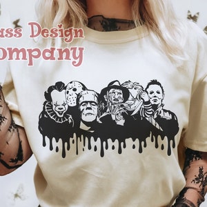 May include: Cream-colored t-shirt featuring a black graphic of iconic horror movie characters, including Pennywise, Jason Voorhees, and Michael Myers. The graphic has a dripping effect at the bottom. The text "Class Design Company" is in the upper left corner.