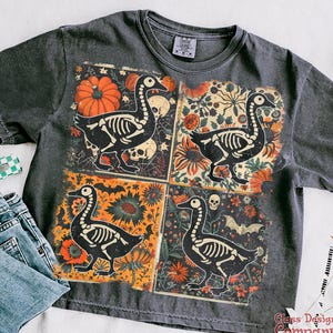 May include: Dark grey t-shirt with a four-panel design featuring a skeleton goose illustration. Each panel has a different background with pumpkins, flowers, and bats. The shirt has a vintage, distressed appearance.