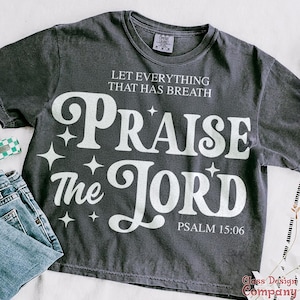 Può includere: Una maglietta grigia scura con una grafica bianca che dice "Che tutto ciò che ha respiro lodi il Signore Salmo 15:06".