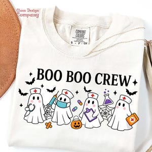 May include: Cream-colored t-shirt with the text "BOO BOO CREW" and illustrations of ghost characters dressed as medical professionals. The design includes bats, spiderwebs, and a jack-o'-lantern, perfect for Halloween.