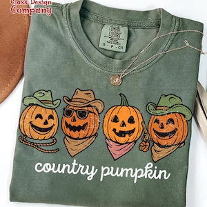 May include: Green Comfort Colors t-shirt featuring a graphic of four orange jack-o'-lanterns wearing cowboy hats and bandanas. The text "country pumpkin" is printed below the pumpkins. A silver necklace is draped across the shirt.