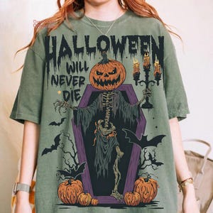 Może przedstawiać: Zielona koszulka z motywem Halloween. Projekt przedstawia szkielet z głową dyni wychodzący z trumny, trzymający świecznik. Tekst "HALLOWEEN WILL NEVER DIE" jest wydrukowany nad postacią.