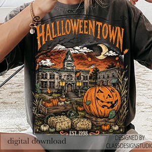May include: A charcoal gray t-shirt featuring a graphic print of a spooky town scene with the text HALLLOWEENTOWN and EST. 1998. The illustration includes a large jack-o'-lantern, a wooden wagon filled with pumpkins, hay bales, and a crescent moon.