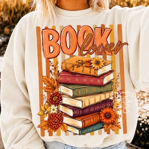 Pode incluir: Uma camisola de gola redonda cor de creme com uma impressão gráfica de uma pilha de seis livros. O texto "BOOK lover" aparece em letras cor de laranja e castanhas sobre os livros. O design inclui flores e folhas de outono em tons alaranjados.