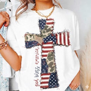 May include: White t-shirt featuring a cross design made of patchwork fabric with American flag and camouflage patterns. The words "god bless america" are written vertically on the left side of the cross. The shirt has a round neckline.