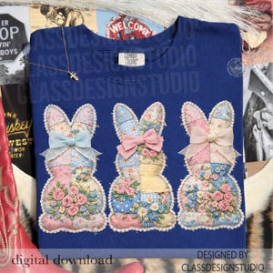 May include: A navy blue t-shirt featuring three appliqued bunny rabbits. Each bunny is made of patchwork fabric with floral and gingham patterns, adorned with bows and roses. The text "CLASSDESIGNSTUDIO" is visible.